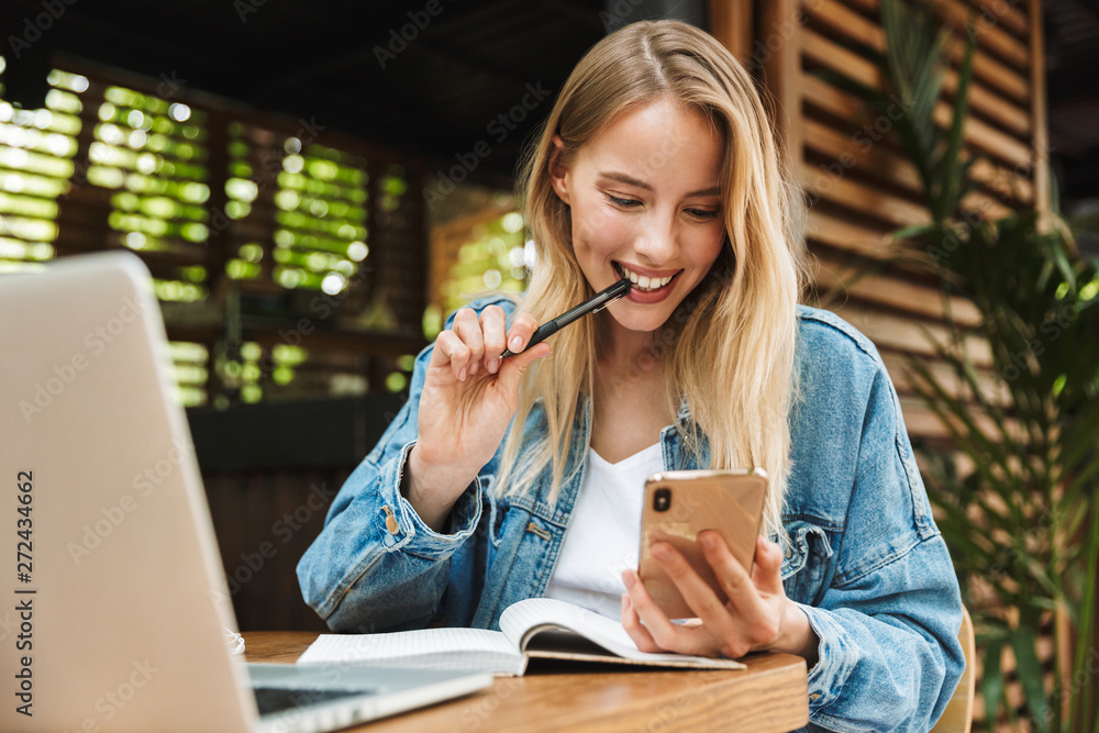 Sticker portrait of caucasian smiling woman writing in diary while using laptop and cellphone in cafe outdoo