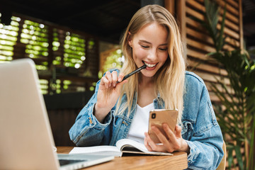 Portrait of caucasian smiling woman writing in diary while using laptop and cellphone in cafe outdoors