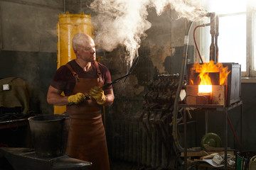 Busy handsome young man blowing on handmade knife held in tongs while examining it after forging and cooling