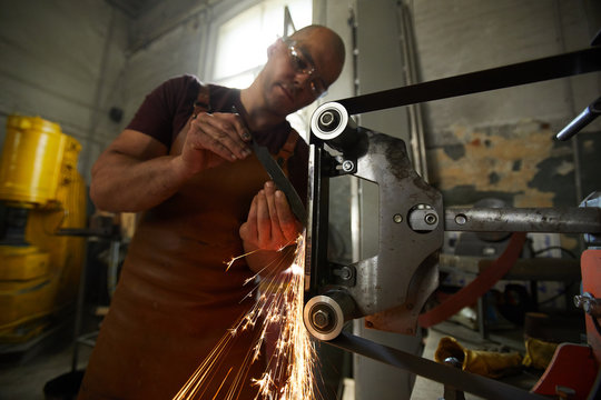 Concentrated young blacksmith in apron standing at grinding machine and sharpening knife in workshop