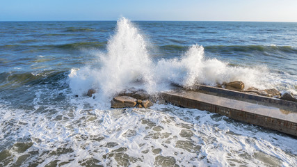 Aerial view on a stone pier against which the force of the sea is raging with high and strong waves. On the construction in the middle of the water there is nobody.Waves banging create the white foam.