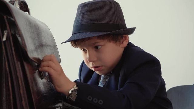 Little boy looks like a boss, in suit and hat with watches on hands. He is trying close his briefcase sitting in his office. Fail boss can not to close briefcase