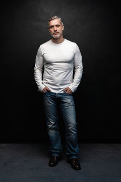 Full body portrait of middle-aged good looking man in white t-shirt posing in front of a black background with copy space.