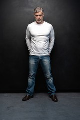 Full body portrait of middle-aged good looking man in white t-shirt posing in front of a black background with copy space.