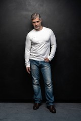 Obraz premium Full body portrait of middle-aged good looking man in white t-shirt posing in front of a black background with copy space.