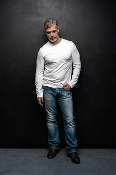 Full body portrait of middle-aged good looking man in white t-shirt posing in front of a black background with copy space.