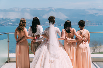 Backside view bride in white dress and girlfriends hold hands on balcony