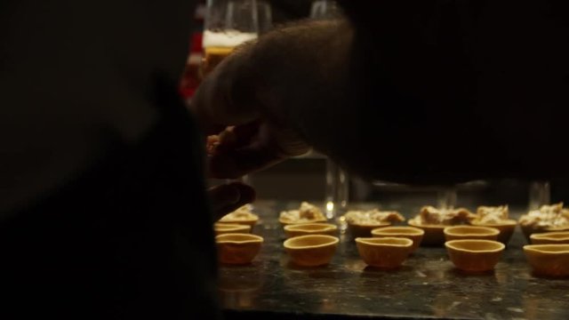 Close-up of someone stuff some small pastry shells in slow motion with glasses of champaign in the background.