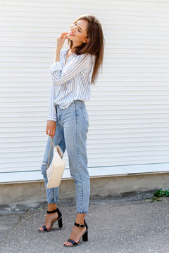 Young stylish woman wearing blue striped shirt, blue cropped denim jeans, black high heel sandals and beige handbag posing outdoors against white street wall. Trendy casual outfit. Street fashion.