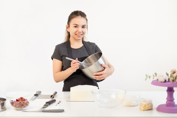 Attractive brunette girl pastry chef in apron whips cream cake in a metal bowl.