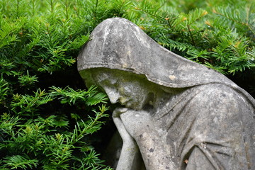 A weathered statue of a sad angel that is surrounded with by branches on a christian cemetery in Berlin-Germany.