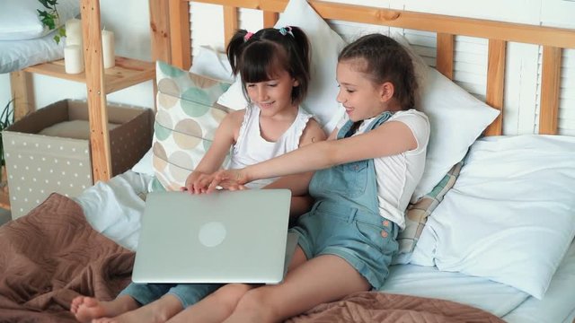 Two little girls sit on bed, open laptop to see cartoons, slow motion