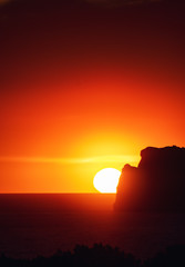 Dramatic ocean sunset scene with giant big sun setting behind a rock coastline silhouette an colorful sky glow. Port Andratx, Mallorca, Spain