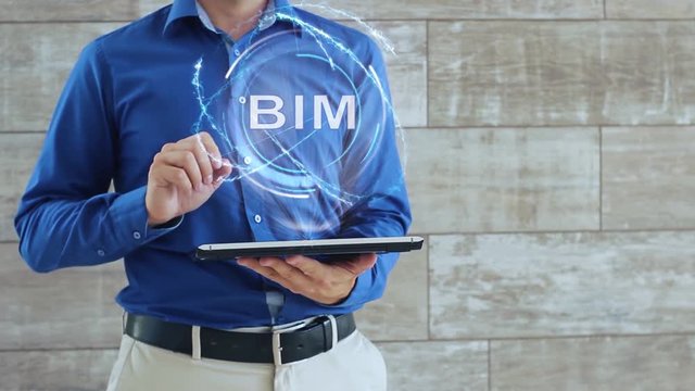 Man activates a conceptual hologram with text BIM. The guy in the blue shirt and light trousers with a holographic screen gadget on the background of the wall