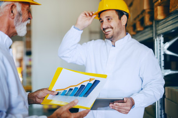 Two male Caucasian employees in white uniforms and with yellow helmets on heads talking about sale growth while standing in warehouse. Younger one holding tablet while older one holding chart.