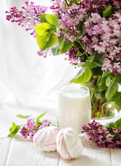 The glass of milk served with handmade marshmallows and a bouquet of a lilac on white table. Breakfast concept. Selective focus. 