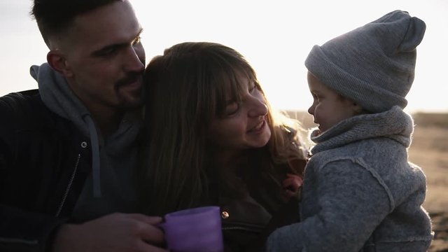 Slow motion of sweet, young family in casualwear sitting on plaid embracing and enjoying time together, drinking hot drinks from plastic cups. Mother touching her child nose, smiling. Cold weather