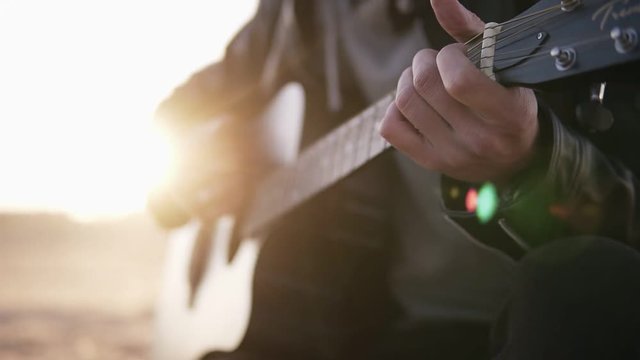 Young caucasian man, stylish bearded standing outdoors on the autumn beach and playing guitar at amazing evening with lens flare effects. Slow motion. Close up