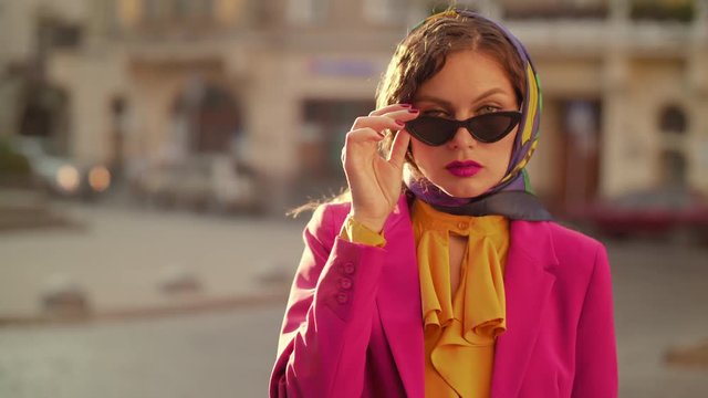Fashion model wearing black cat eye sunglasses, kerchief, colorful clothes, looking at camera, posing in street of european city