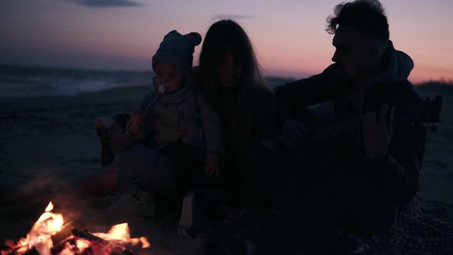Adorable, young family together with their daughter on the beach with a fire and a guitar, the concept of family values, love and closeness