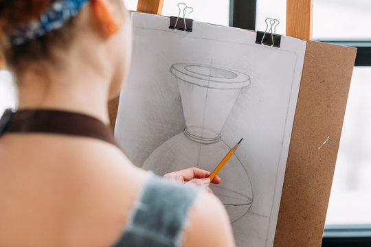 Art studio. Closeup back view of lefthanded painter measuring proportions with pencil in drawing vase.