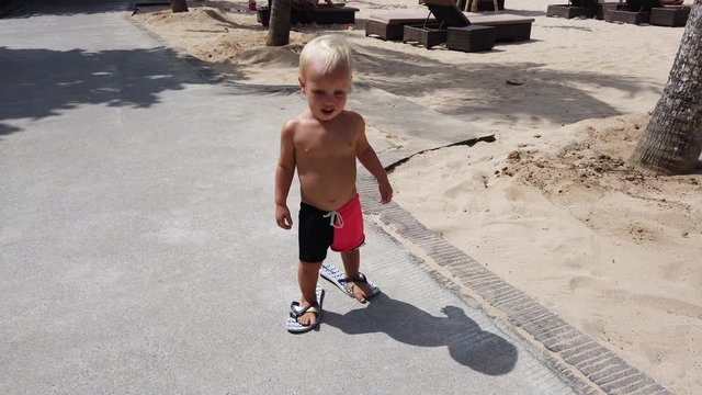 Cute two year old white boy trying to walk down the path in big female summer flip flops. beach in the background.