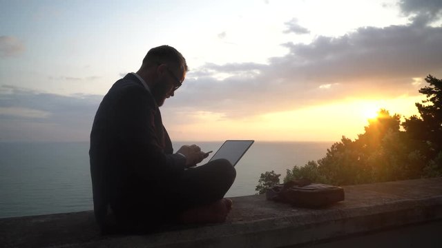 young businessman in a suit without shoes is sitting and working in a laptop on a concrete parapet on the background of a sea sunset