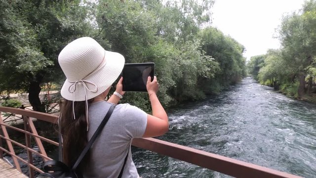 On a wooden bridge across the river girl tourist in hat on his head holding a tablet and takes pictures of the beautiful view of nature