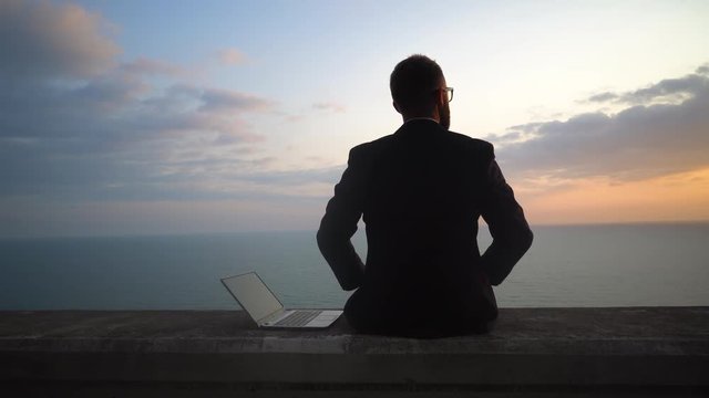 young businessman in a suit without shoes is sitting and working in a laptop on a concrete parapet on the background of a sea sunset