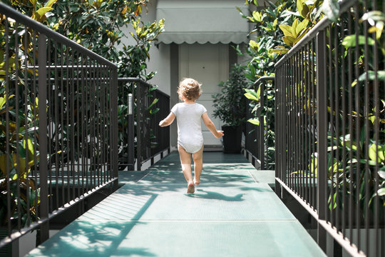 Little Toddler In A White Onesie Running Away From Home To The Courtyard Garden