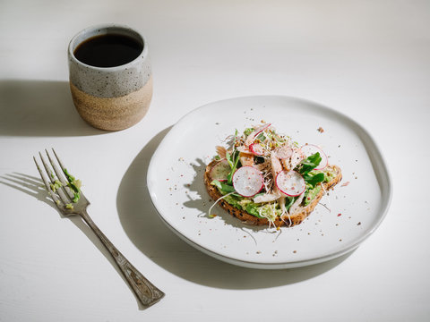 Trendy avocado toast topped with smoked trout fish, arugula, radish, sea salt, and black pepper. Served with coffee in a artisan hand made ceramic mug.