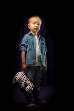 A Little Boy In A Toddler Denim Shirt Is Standing And Holding A Soft Toy Zebra In His Hand. Full-length Portrait On A Black Background.