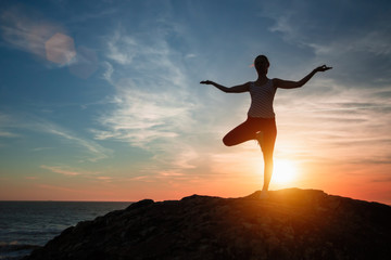 Silhouette of woman standing at yoga pose on the sea beach during sunset.