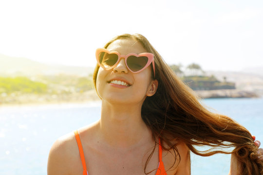 Pretty brunette woman in pink heart sunglasses on the beach. Holidays, vacation travel and freedom concept. Beautiful happy girl in heart shaped sunglasses on beach.