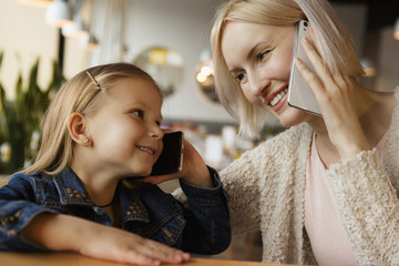 View from side of happy cute family sitting in cafe and talking by phone. Pretty girl in jeans jacket and attractive mother looking at each other and laughing. Concept of happiness and relaxing.