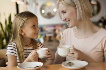Side view of attractive mother drinking green tea with little daughter in cafe. Smiling girl and woman keeping cups in hands, looking at each other and enjoying family time. Concept of coffee.