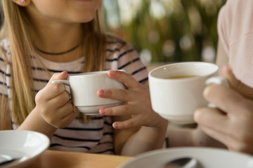 Selective focus of cups of green tea in hands of little girl and woman in cafe. Daughter and mother drinking cappuccino and coffee during lunch, talking and smiling. Concept of coffee and enjoyment.