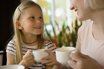 Cute girl keeping cup of warm tea, looking at mother and smiling in cafe. Lovely family sitting at table having lunch, drinking cappuccino and talking in restaurant. Concept of relaxation.