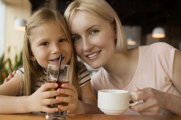 Front view of cheerful mother and daughter looking at camera and smiling while drinking juice and coffee in cafe. Lovely family posing while having rest together. Concept of lunch and love.
