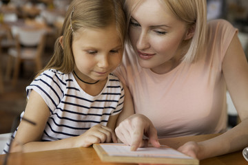 Front view of cute mother and daughter sitting in cafe, looking at menu and choosing meal. Lovely...