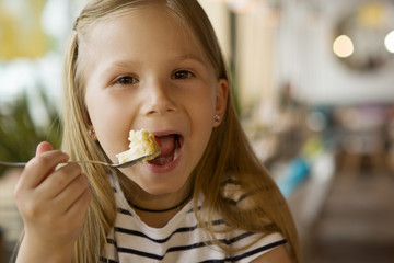 Happy girl looking at camera while eating tasty cake in cafe. Pretty kid with open mouth keeping fork, smiling and posing. Funny child relaxing and resting on weekends. Concept of food and joy.