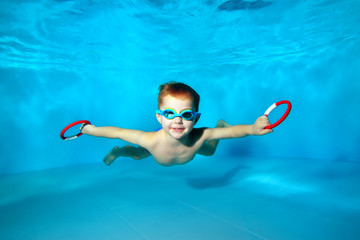 Sporty little boy swimming underwater in the pool with toys in his hands. Portrait. Close up. Underwater photography. Horizontal orientation - obrazy, fototapety, plakaty