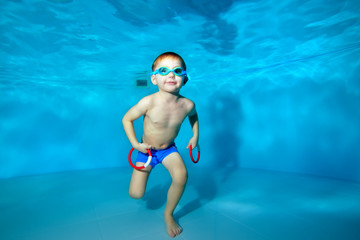 Sporty little boy standing under water at the bottom of the pool with toys in his hands. Swimming...