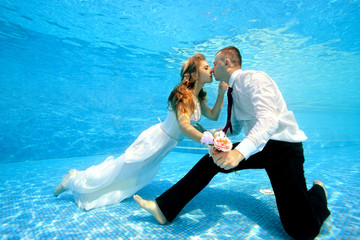 Happy bride and groom hold hands and kiss underwater at the bottom of the pool on a Sunny day. Wedding bouquet in hand. Portrait. Concept. Underwater photography. Horizontal view