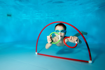 Sporty little boy swimming underwater at the bottom of the swimming pool with toys in his hands....