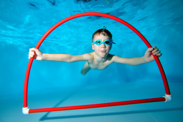 A little boy is engaged in swimming in the pool, underwater at the bottom, posing with a red Hoop...