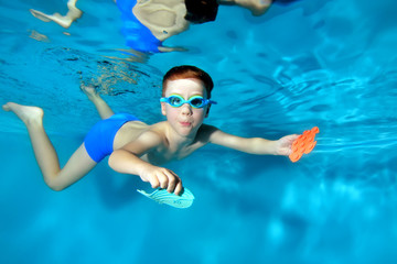 Little sports boy swims underwater,dives to the bottom of the pool with toys in . Posing for the camera with his eyes open. Portrait. Photo underwater. Horizontal orientation - obrazy, fototapety, plakaty