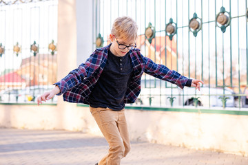 Funny blonde boy in gray hat learning to skate on black skateboard in the warm green park in the centre of big city. Summer activity concept