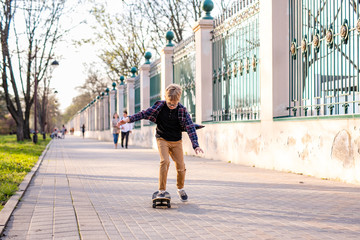 Funny blonde boy in checkered shirt learning to skate on black skateboard in the warm green park in the centre of big city. Summer activity concept