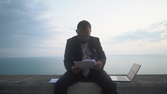 young businessman in a suit without shoes is sitting and working in a laptop on a concrete parapet on the background of a sea sunset